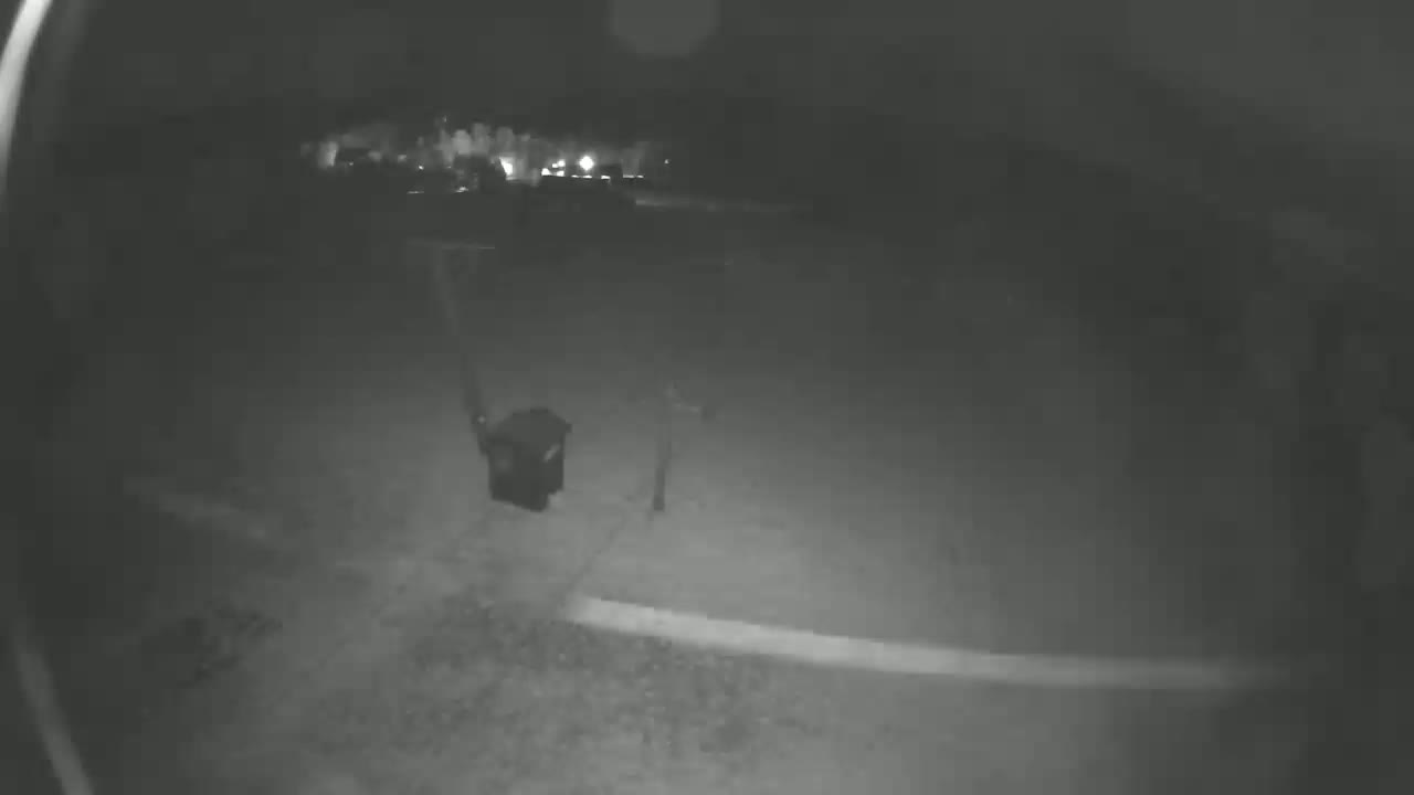 A nighttime, low-light image shows a blurry view of a residential area with a dark-colored trash can near a post.
