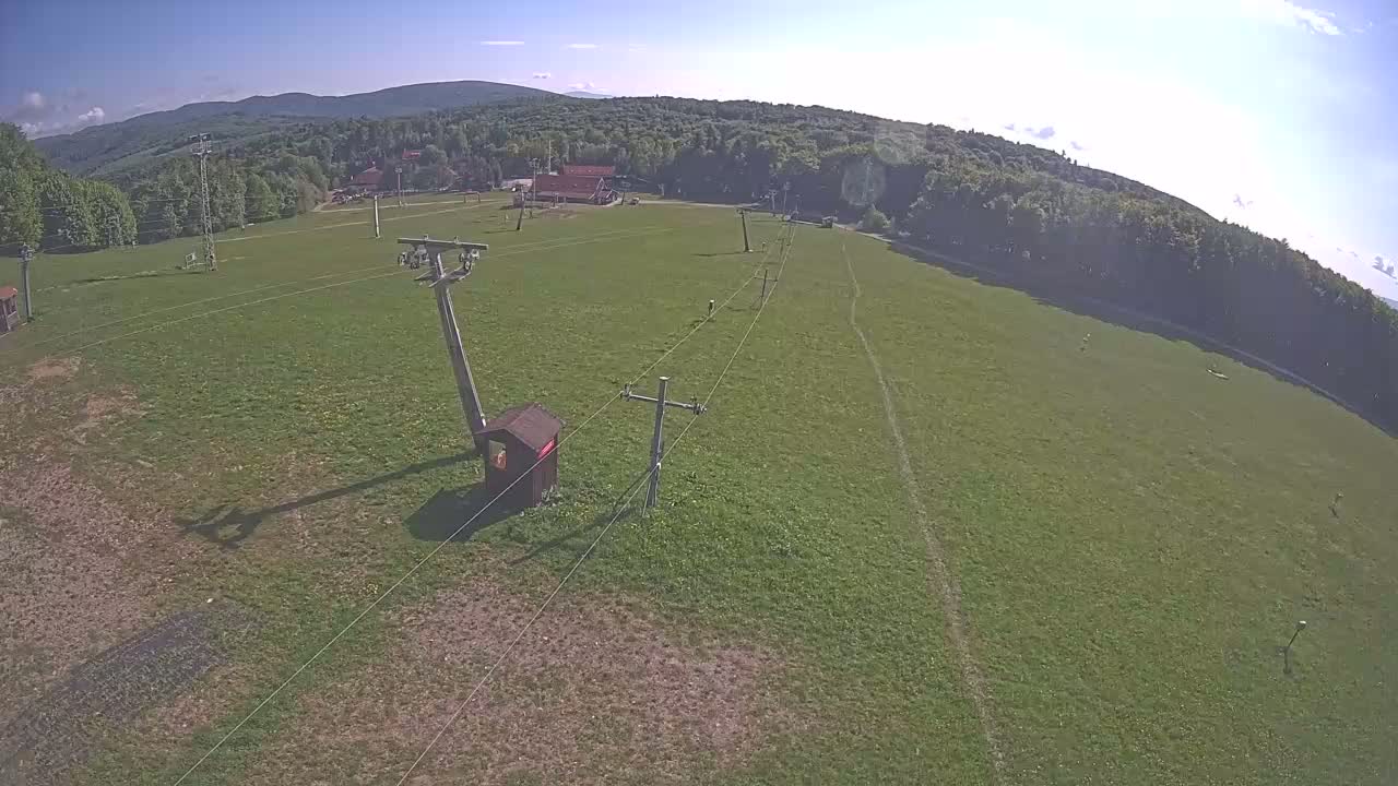 A grassy field with a chairlift and support structures is visible, under a mostly sunny sky, with a line of trees and distant mountains in the background.