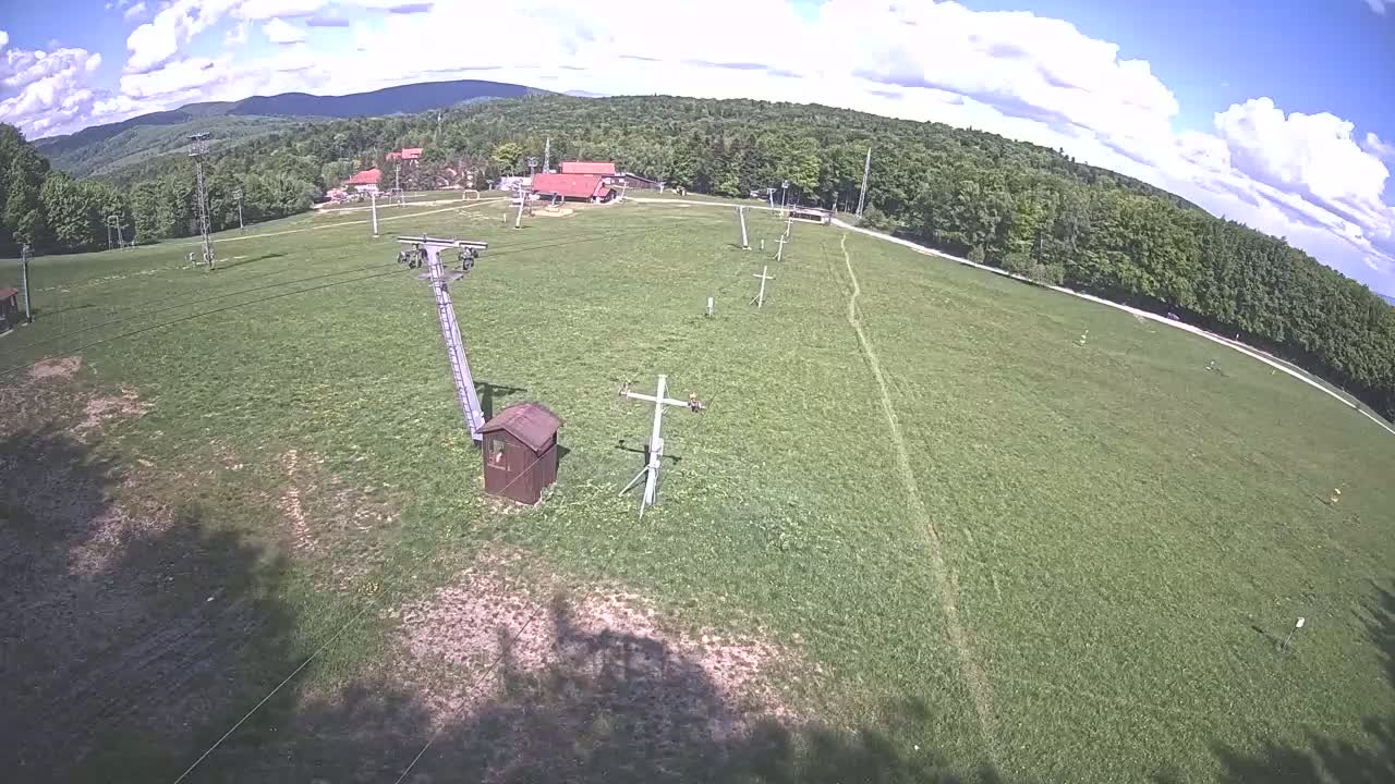 A mostly sunny day shows a grassy field with a chairlift and several support structures, bordered by a forest and distant mountains.