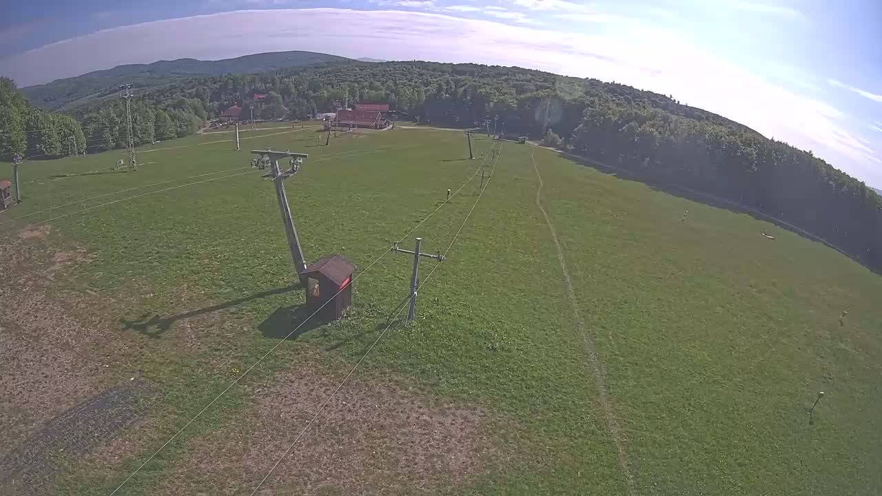 An aerial view shows a grassy field with a ski lift and supporting structures under a mostly sunny sky.