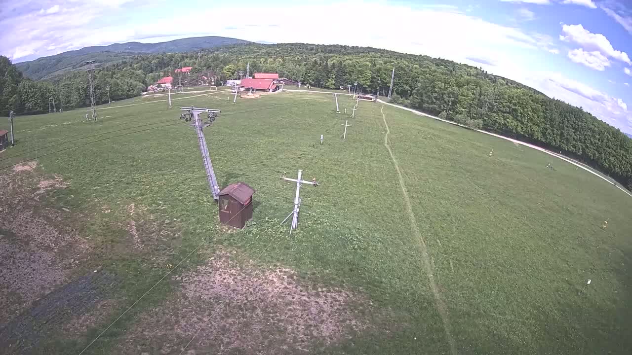 A grassy field with a chairlift, small buildings, and a forest in the background under partly cloudy skies.