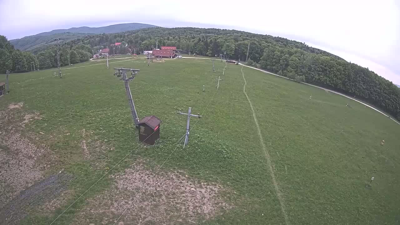 An overhead view shows a grassy field with a chairlift and supporting structures, bordered by a forest under an overcast sky.