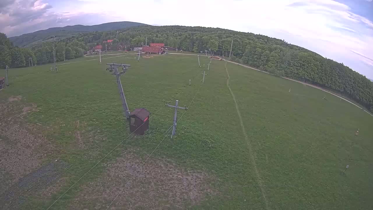 An aerial view shows a grassy field with a ski lift and small buildings under a partly cloudy sky.