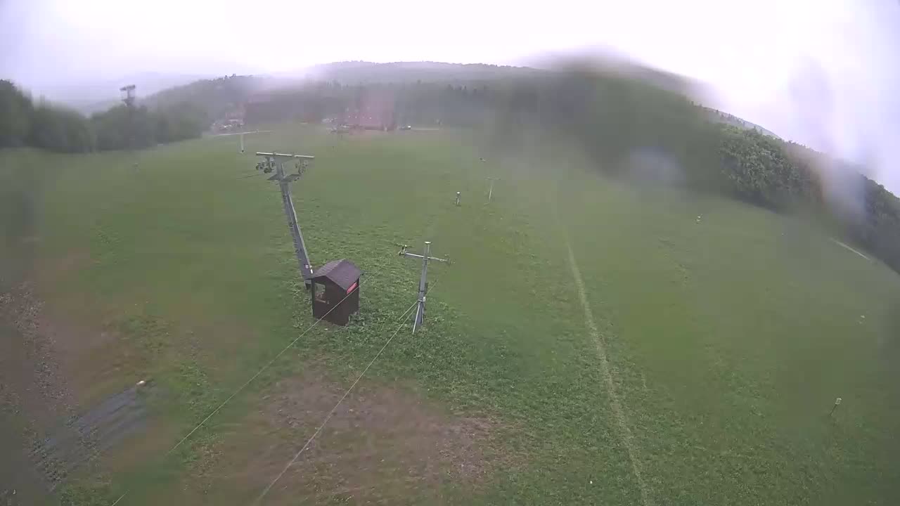 A chairlift and small building are situated on a grassy hill in rainy weather, with a hazy forest in the background.