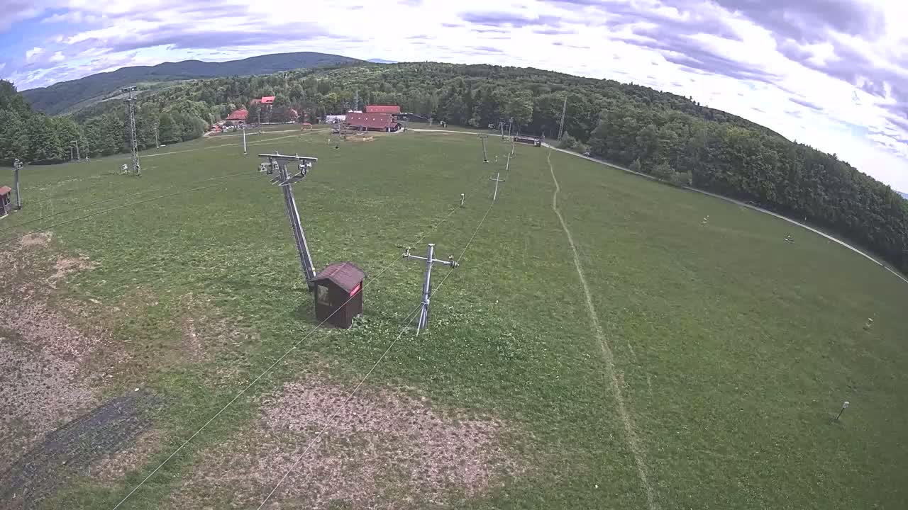An aerial view shows a grassy field with a ski lift and a small building under partly cloudy skies, backed by a line of trees and distant mountains.