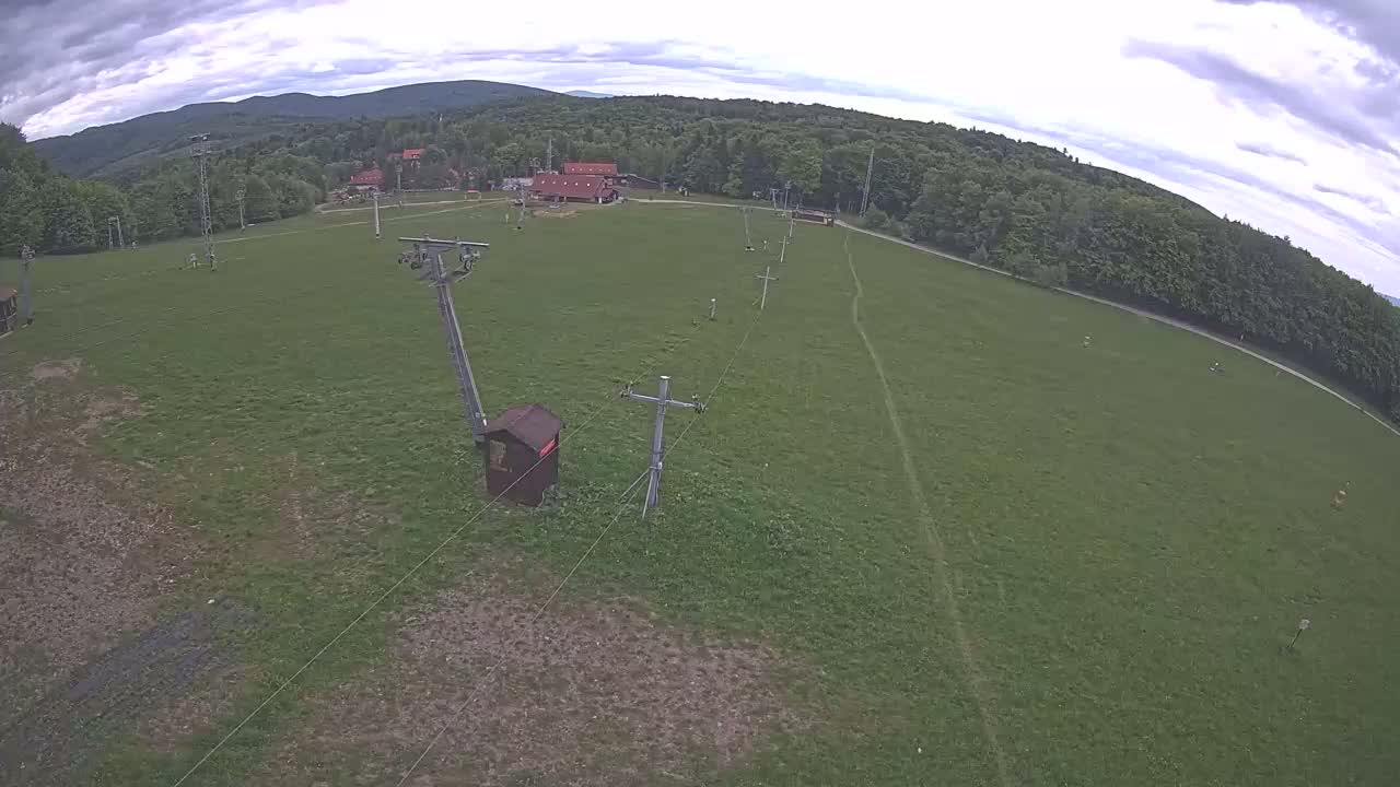 An aerial view shows a grassy field with a chairlift infrastructure, a few small buildings, and a forest-covered mountain in the background under a partly cloudy sky.