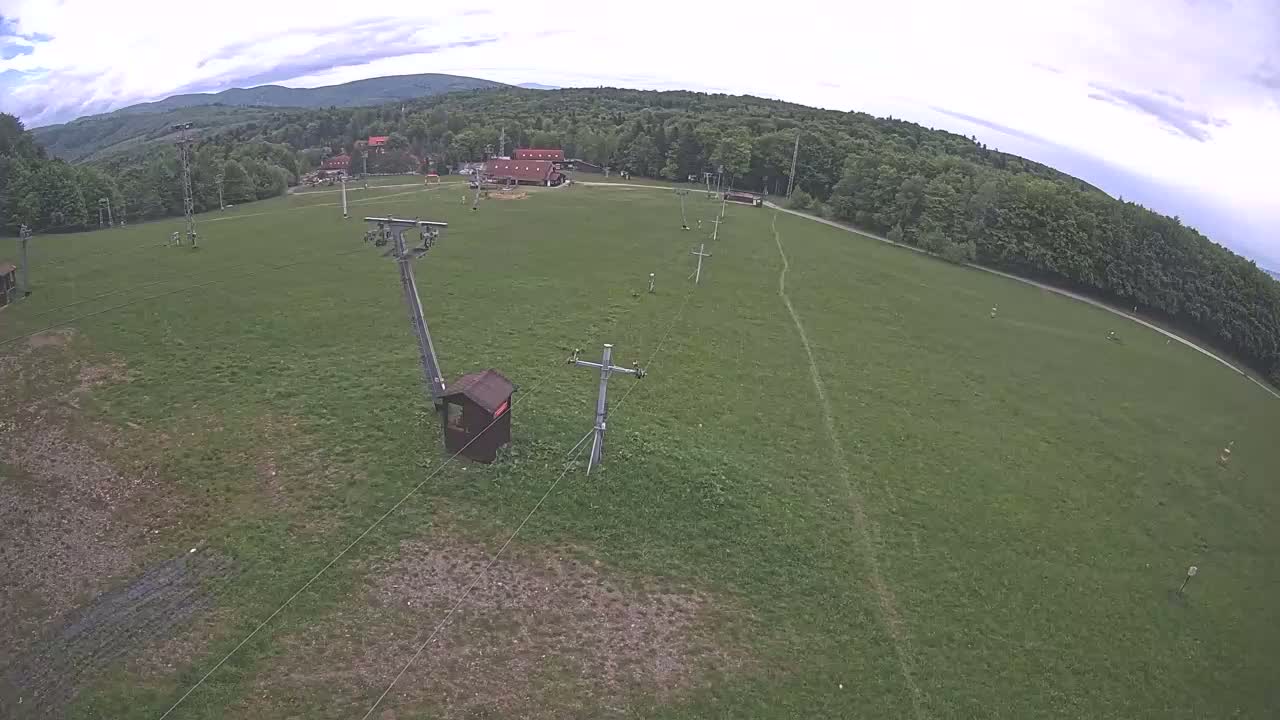 An overhead view shows a grassy field with a ski lift and support structures, under a mostly cloudy sky, surrounded by a forest and distant mountains.