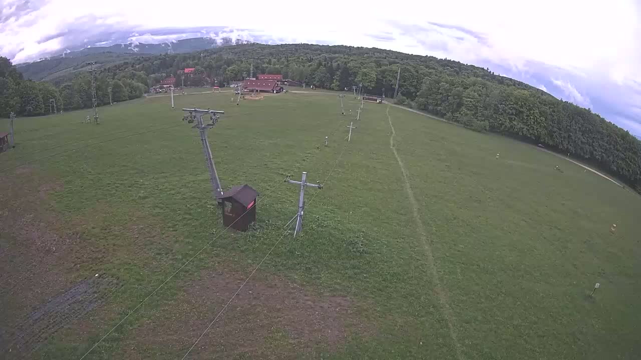 An aerial view shows a grassy field with a chairlift system, a few buildings in the distance, and a mostly cloudy sky.