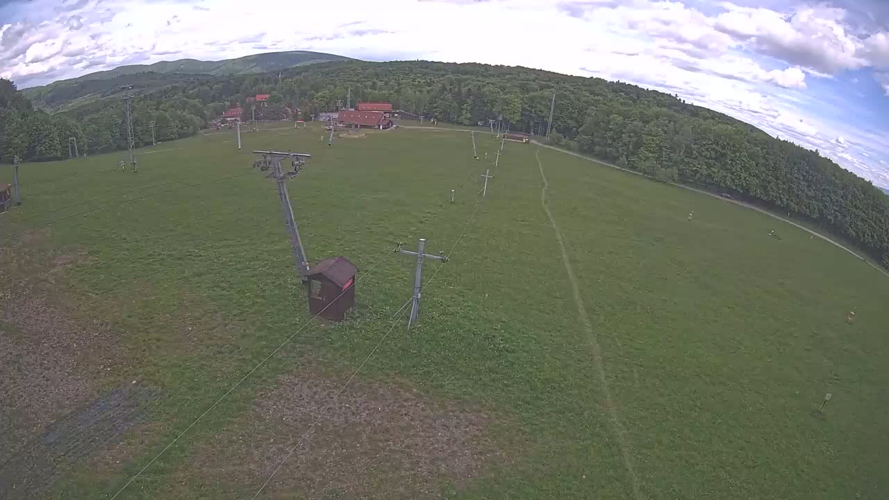 An aerial view shows a grassy field with a chairlift and several buildings nestled in a forest under a partly cloudy sky.