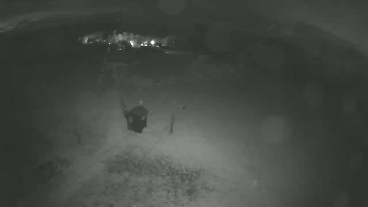 A nighttime, low-light security camera view shows a dark figure near a trash can in a gravel area.