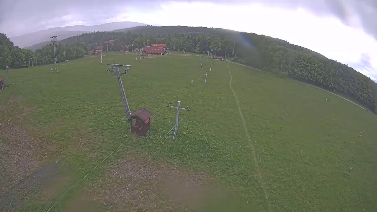 An aerial view shows a grassy field with a ski lift and a small building under an overcast sky.