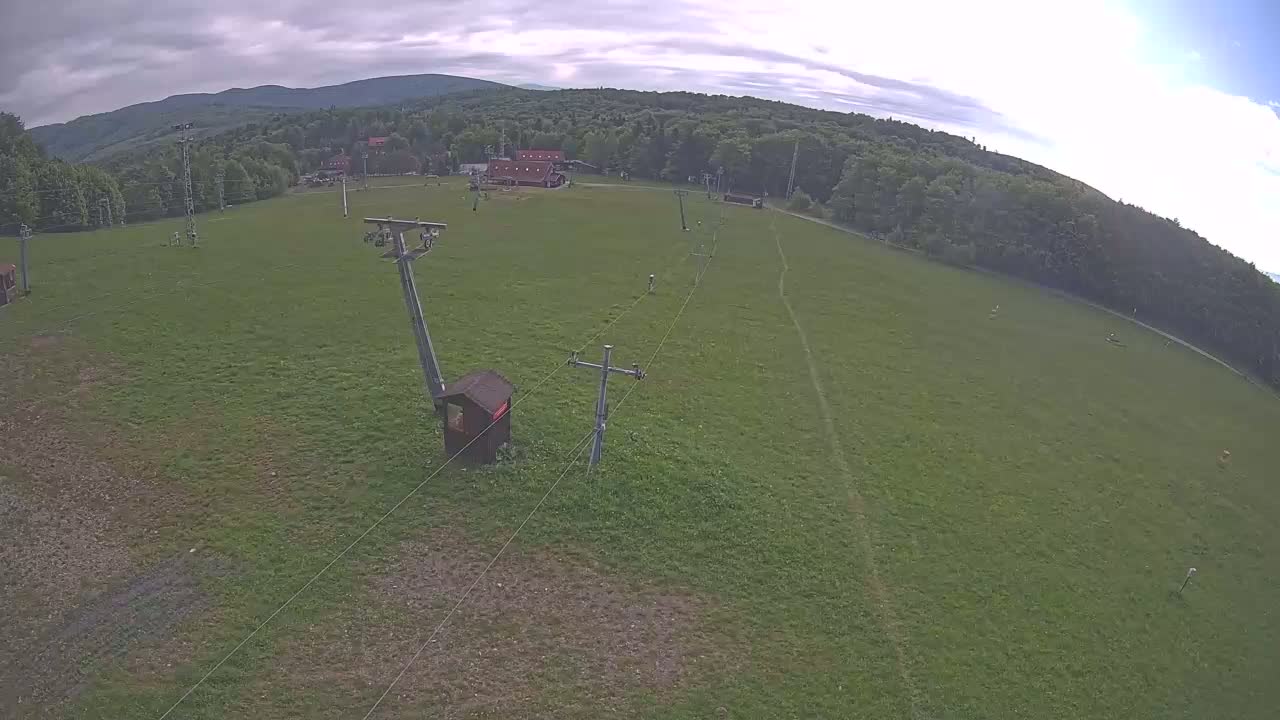 An aerial view shows a grassy field with a ski lift, a small building, and a forested mountain in the background under cloudy skies.
