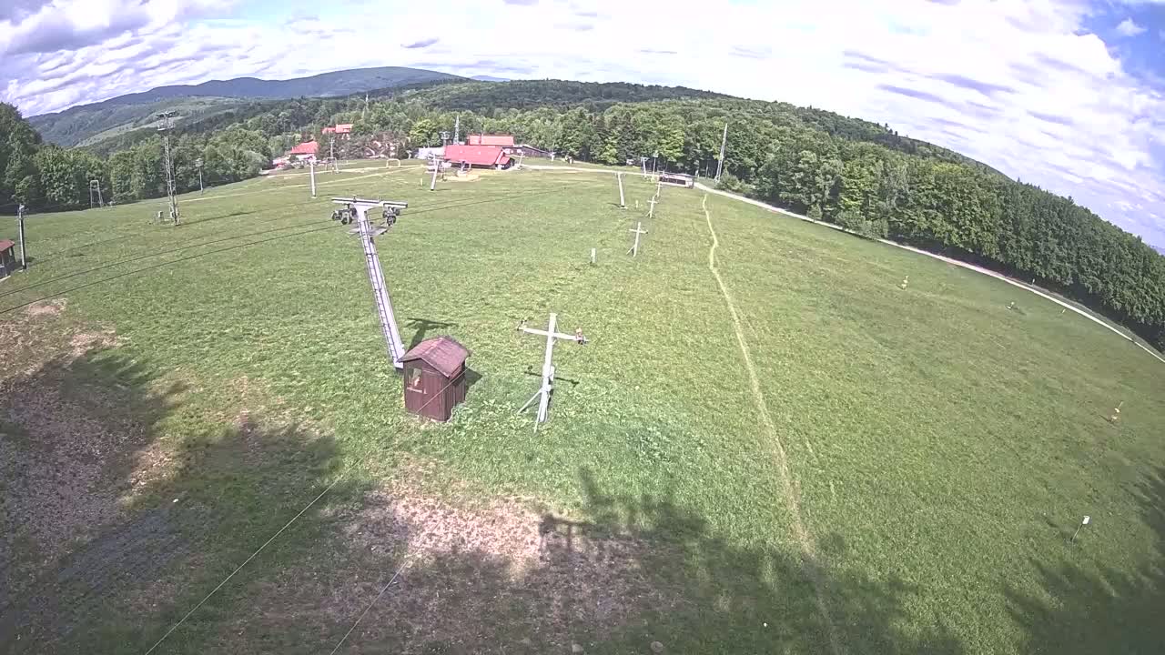 A ski lift sits in a grassy field surrounded by a forest under a partly cloudy sky.