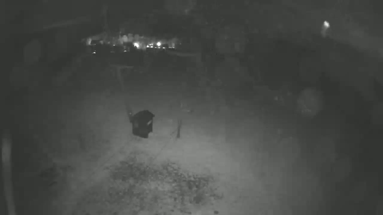 A nighttime, grainy, low-light image shows a dark object, possibly a trash can, in a gravel area, with blurry lights in the background.