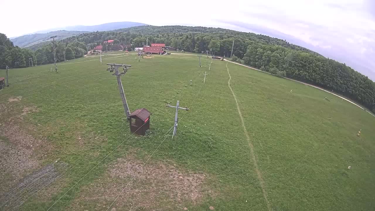 An overhead view shows a grassy field with a chairlift infrastructure, a small wooden building, and a forest in the background under overcast skies.