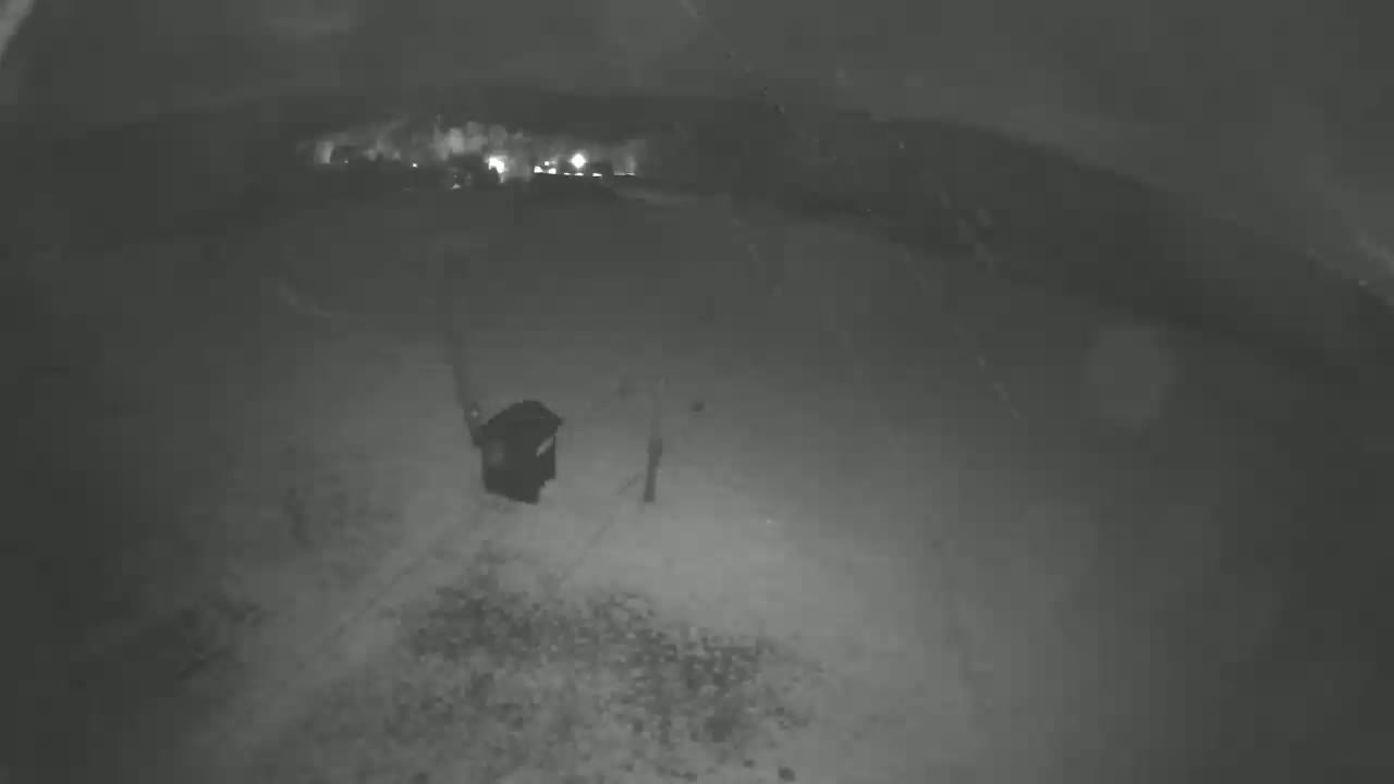 A nighttime, low-light view shows a dark, snow-covered ground with a dark object resembling a trash can near the bottom center, and distant lights visible in the background.