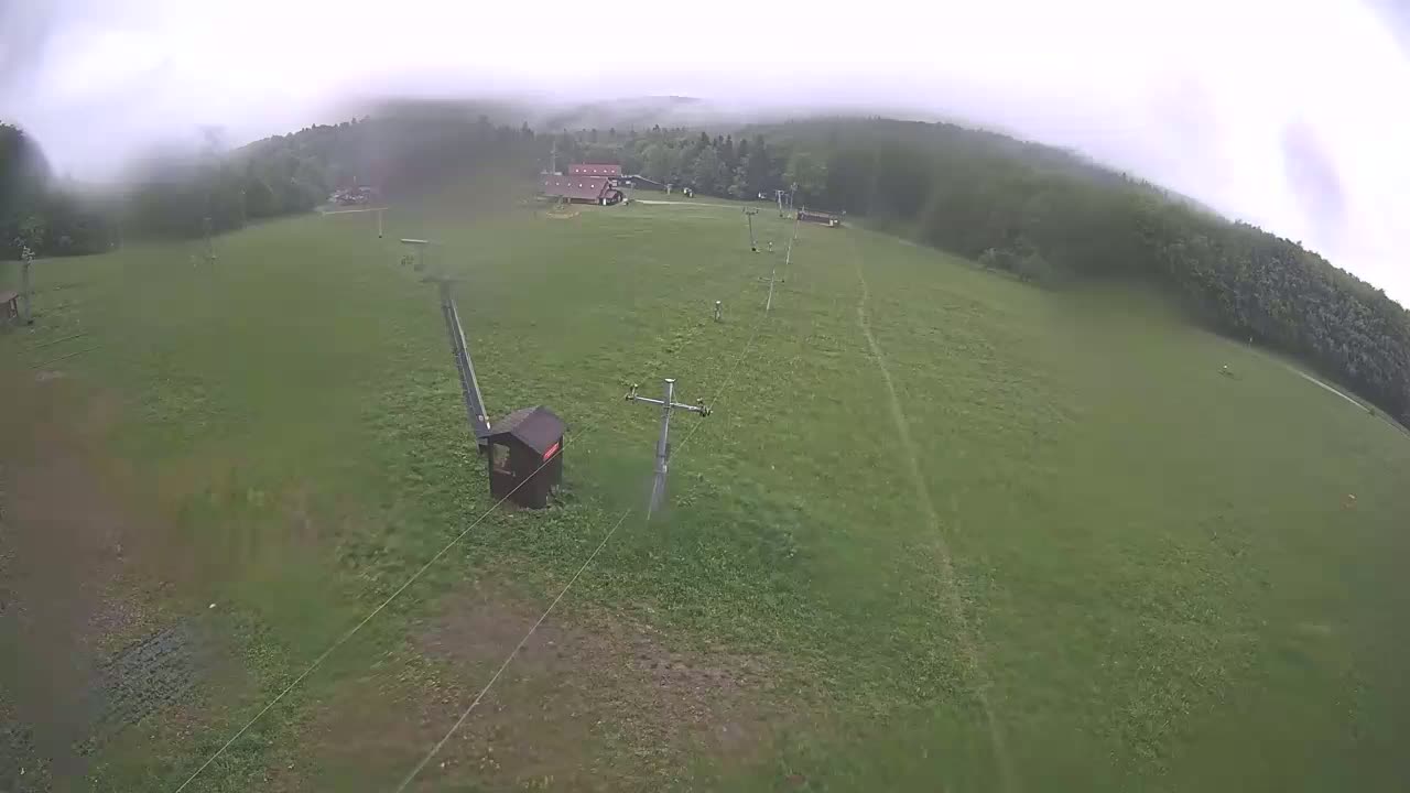 An overhead view shows a grassy field with a ski lift and a small building, under an overcast sky.