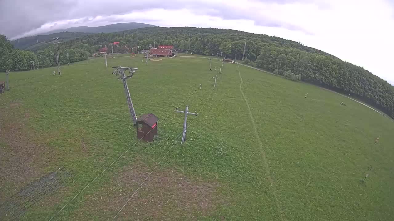 An overhead view shows a grassy field with a ski lift and small buildings under an overcast sky, surrounded by a line of trees and distant mountains.