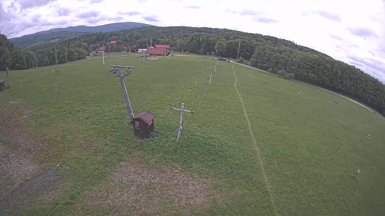 An aerial view shows a grassy field with a ski lift infrastructure, a few buildings, and a forest-covered mountain in the background under cloudy skies.