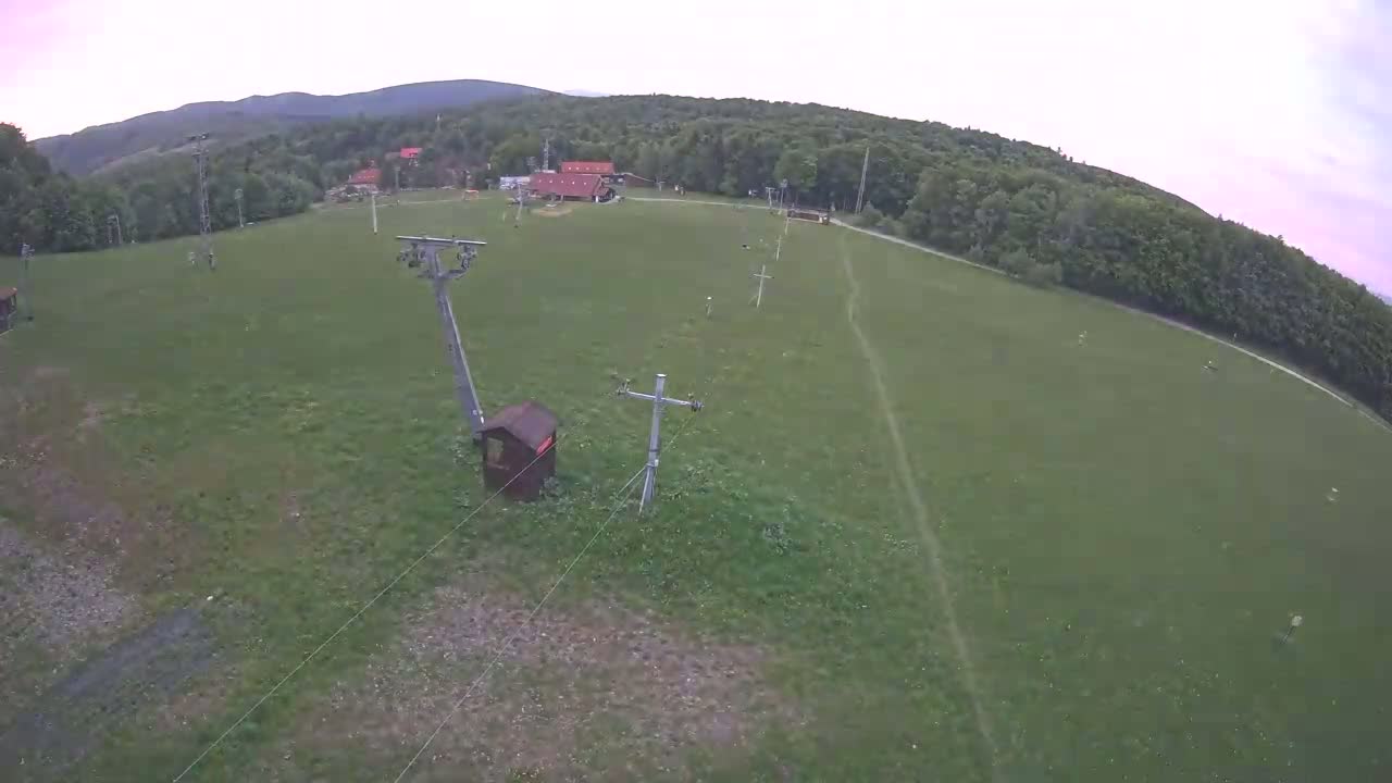 An aerial view shows a grassy field with a ski lift and a small building, under a partly cloudy sky, surrounded by a forest and distant hills.