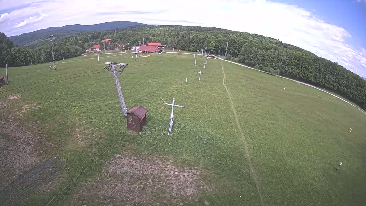 An aerial view shows a grassy field with a ski lift and supporting structures, surrounded by a forest under a partly cloudy sky.