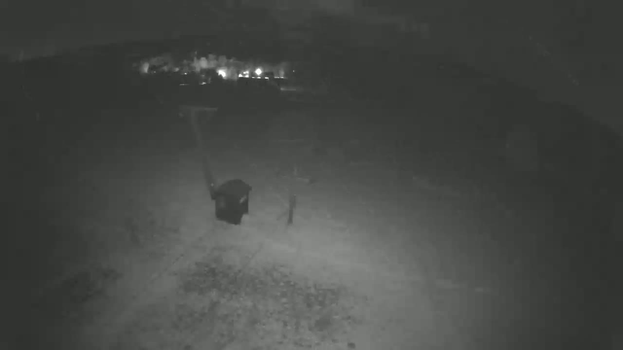 A nighttime, low-light image shows a dark, possibly trash receptacle standing upright near a dimly lit area, under a dark sky.
