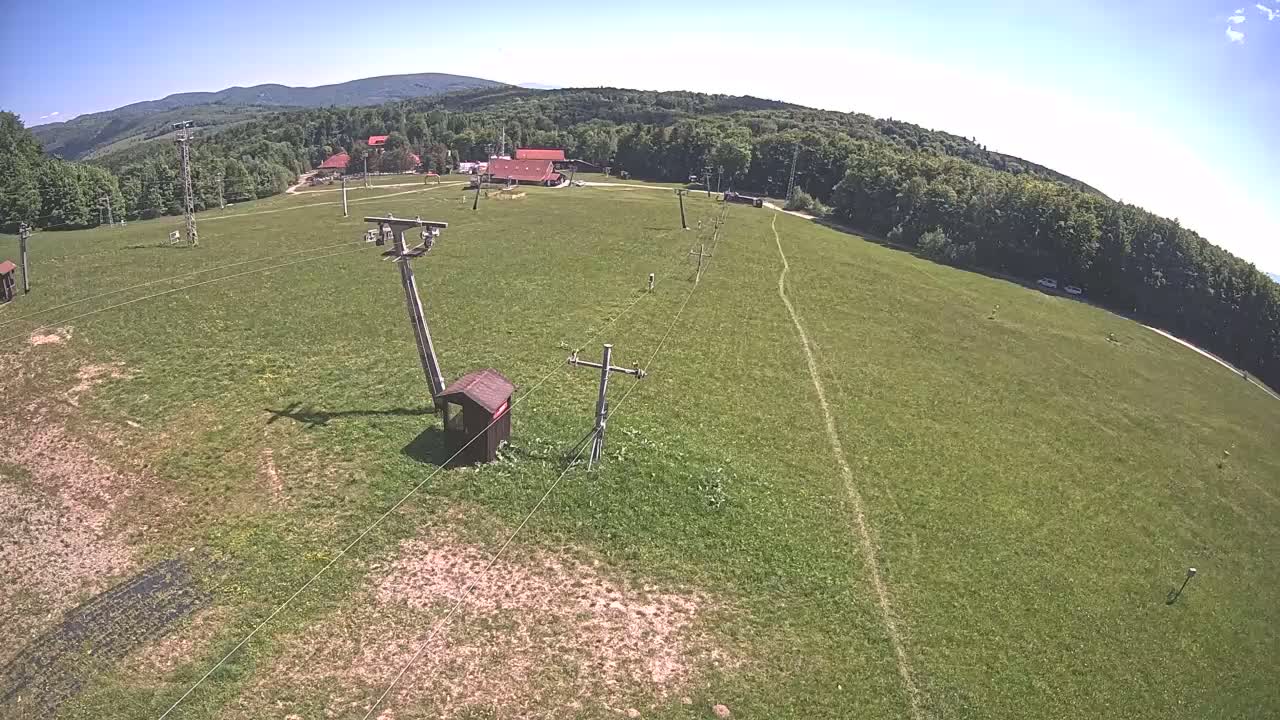 An aerial view shows a grassy ski lift area with a few buildings in the background on a sunny day.