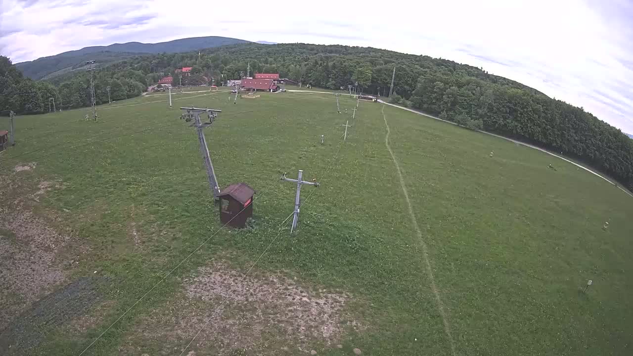 An aerial view shows a grassy field with a ski lift infrastructure and a small building, under a partly cloudy sky, backed by a line of forest and distant mountains.