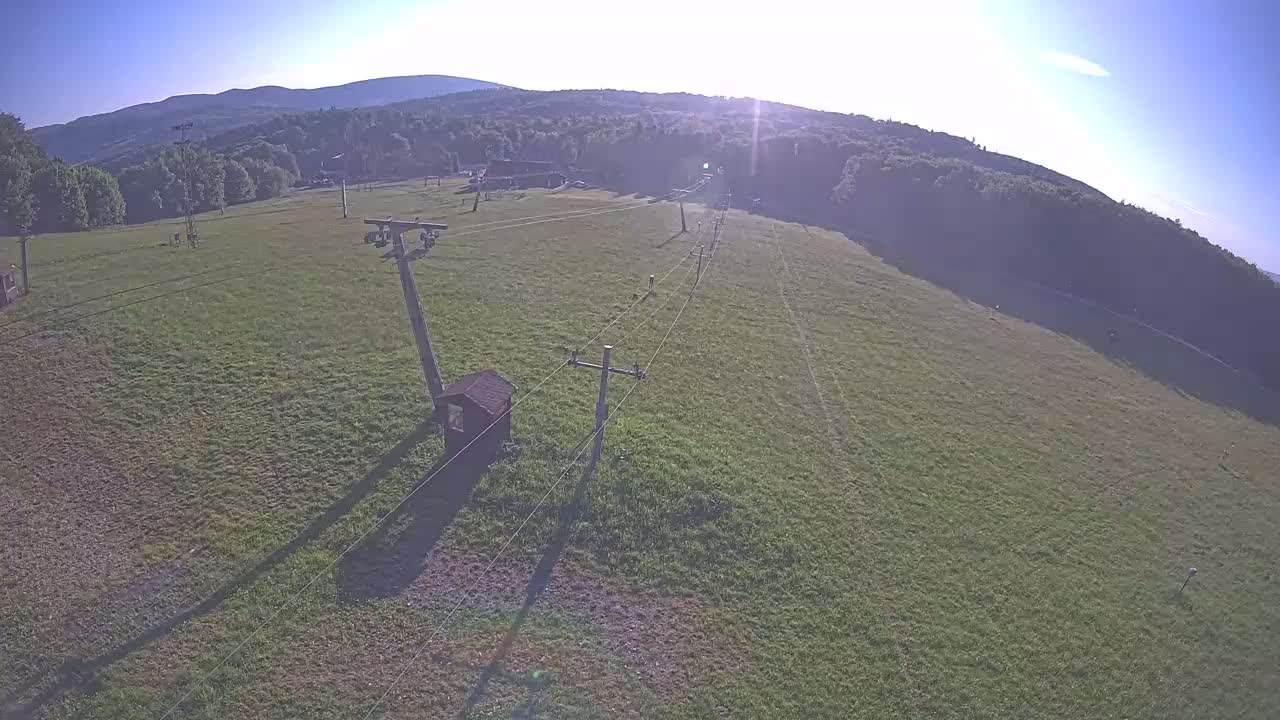 An aerial view shows a grassy field with a ski lift and utility poles under a sunny sky.