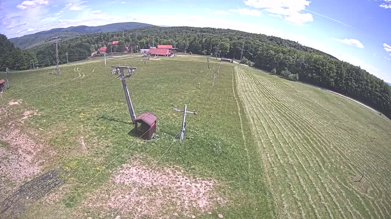 A mostly sunny day reveals a grassy field with a ski lift infrastructure, a small building, and a forested hillside in the background.