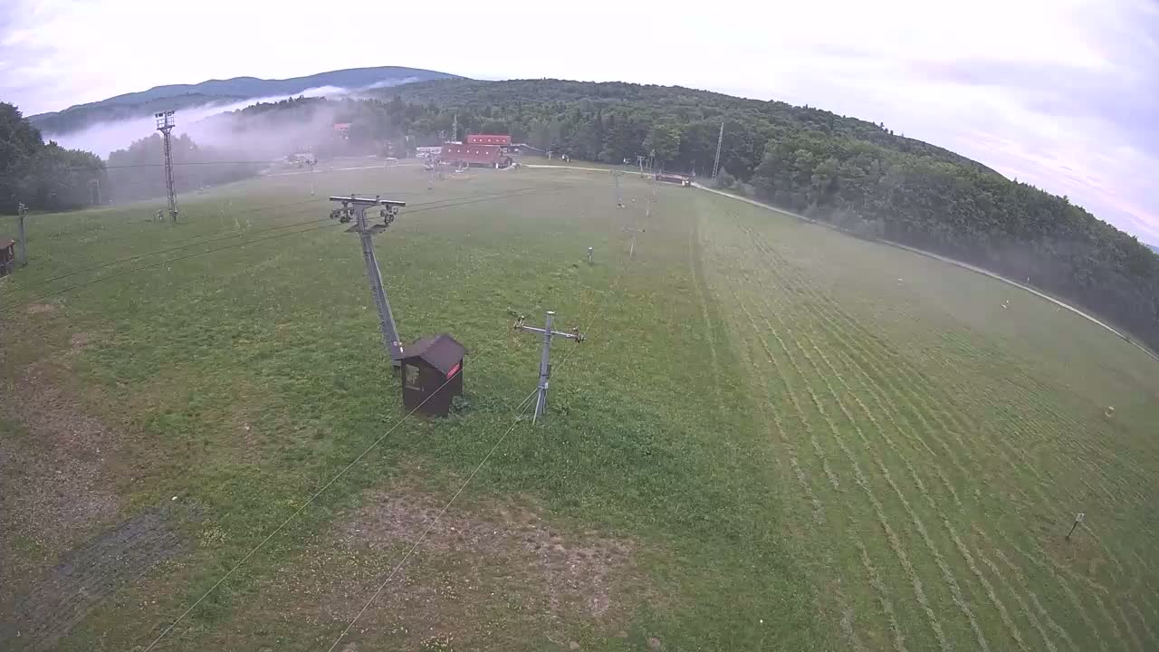 An aerial view shows a grassy field with a chairlift and a building in the distance, under foggy conditions.