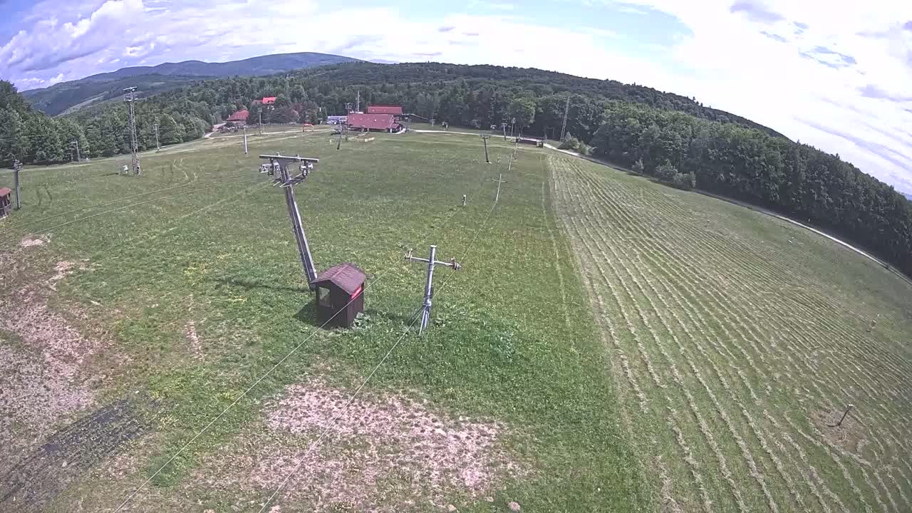 An aerial view shows a grassy field with a recently mowed section, ski lift equipment, and a few buildings in the background under a partly cloudy sky.
