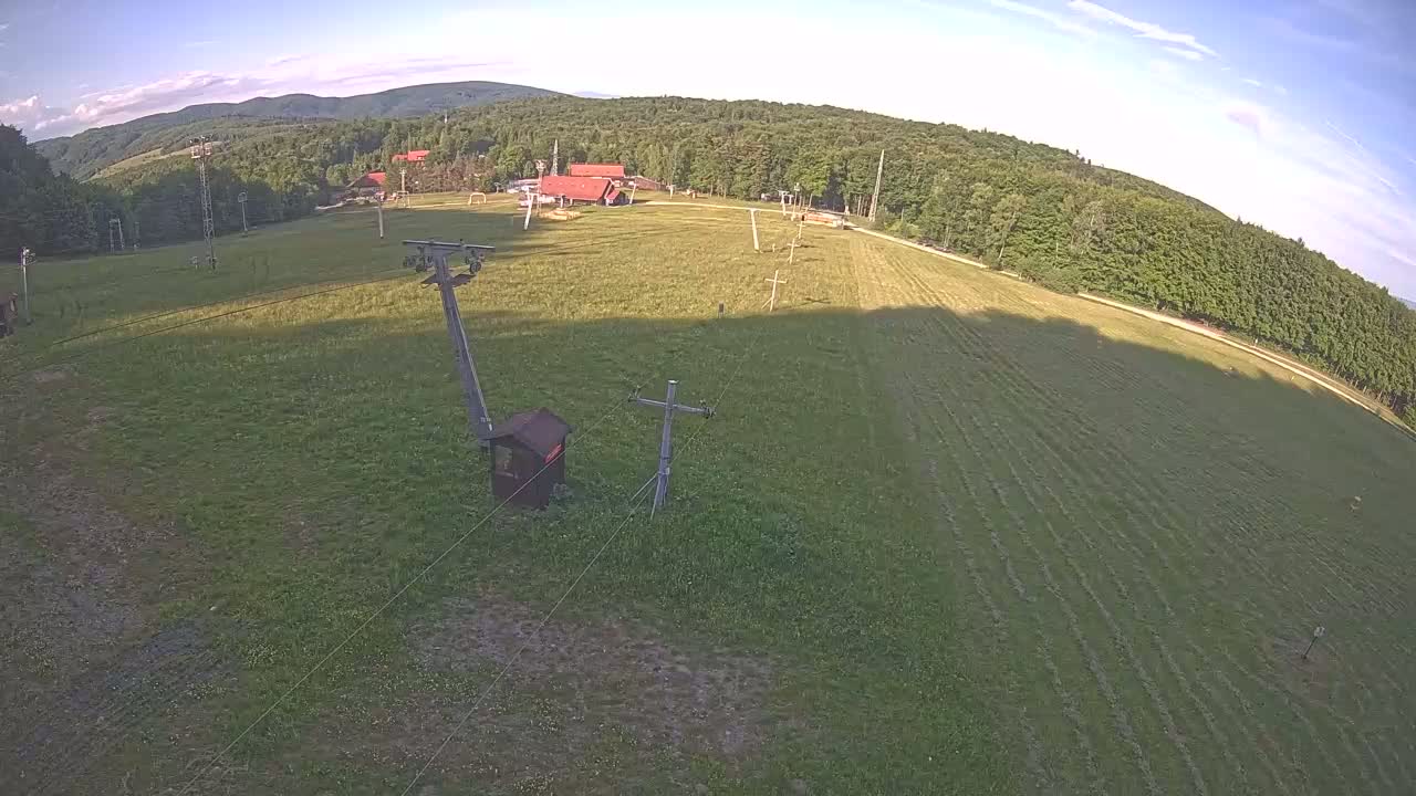 An aerial view shows a grassy field with a ski lift and small buildings, surrounded by a forest under a mostly clear sky.