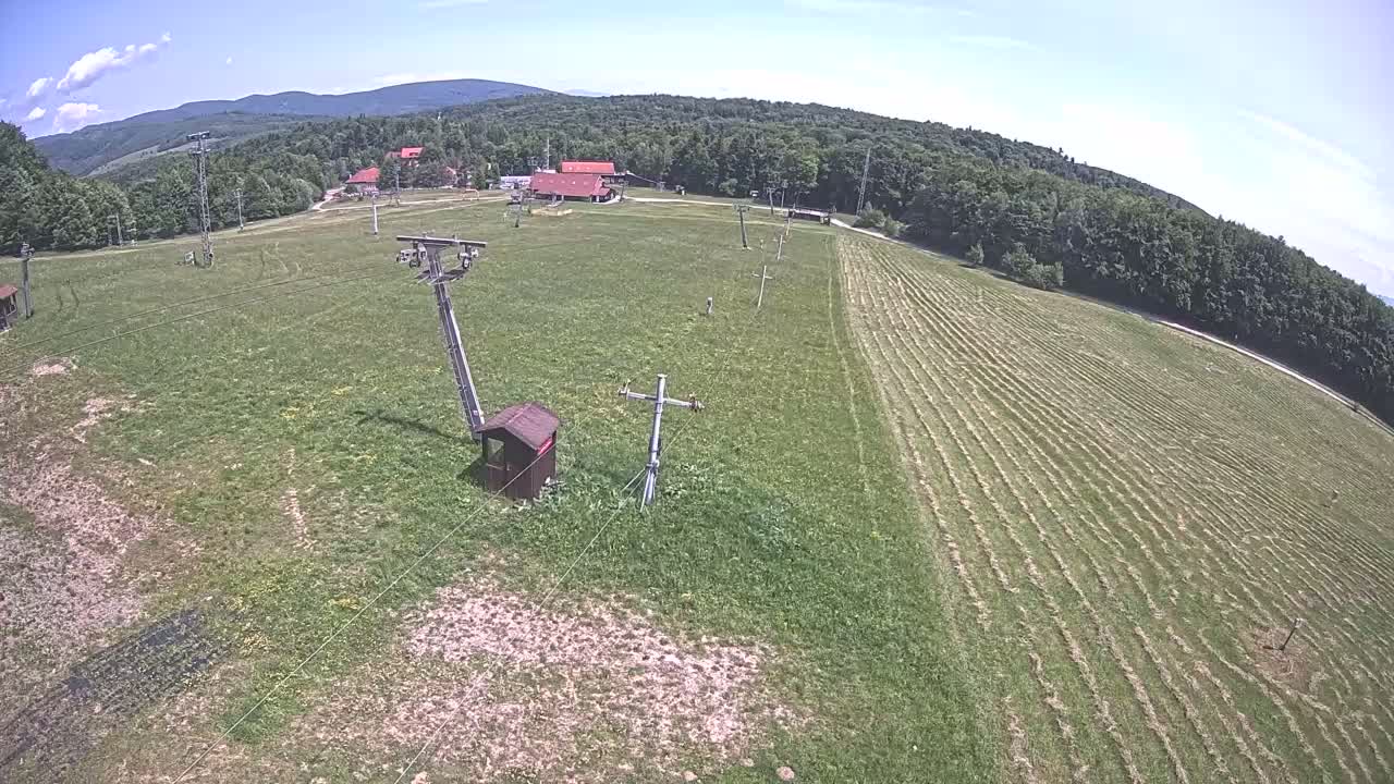 A grassy field with recently cut swaths, ski lift infrastructure, and a small building is visible under a mostly sunny sky, with mountains in the background.