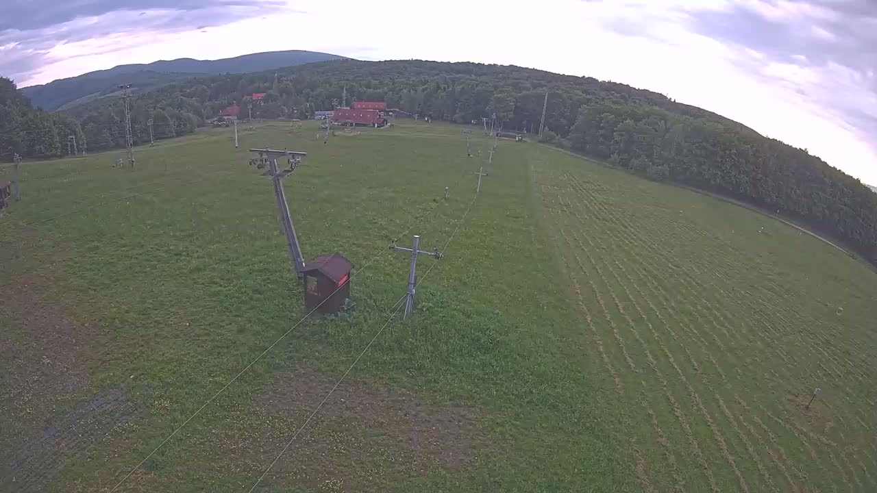 An aerial view shows a grassy field with a ski lift and supporting structures, a few buildings in the distance, and a line of trees beyond under a partly cloudy sky.