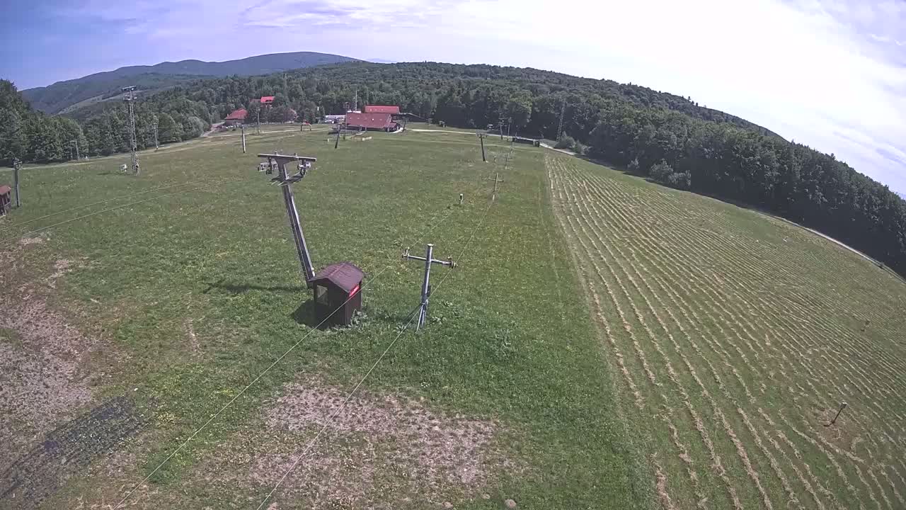 An aerial view shows a grassy field with a ski lift infrastructure, a few buildings, and a forest of trees under a partly sunny sky.