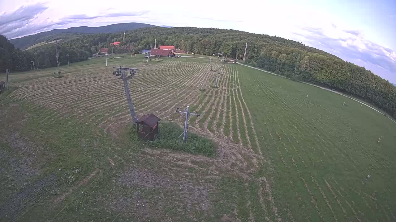 An aerial view shows a grassy field with parallel lines, several ski lift poles, and a small building, under a partly cloudy sky and bordered by a forest and distant mountains.