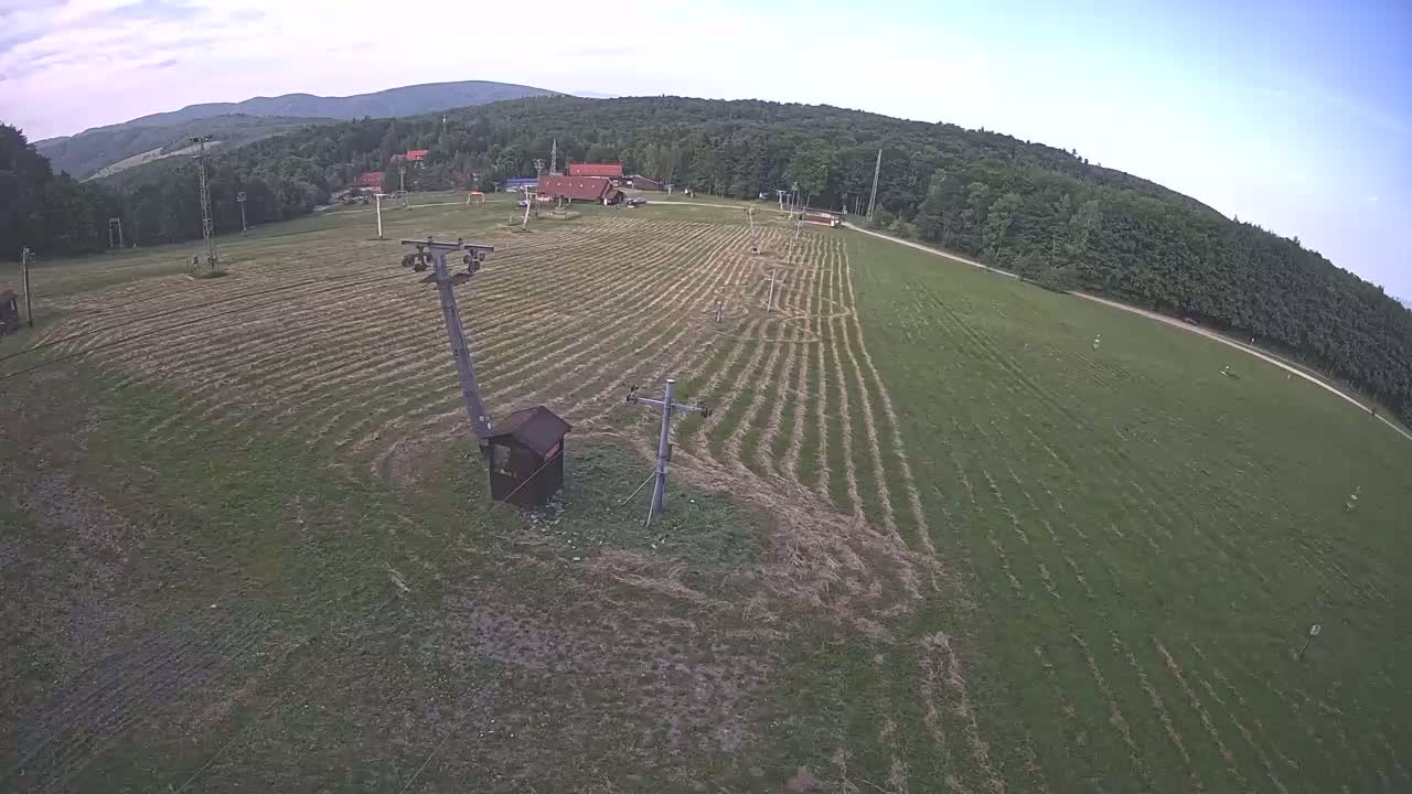 An aerial view shows a grassy field with lines cut into the grass, ski lift infrastructure, a small building, and a wooded hillside in the background under a mostly clear sky.