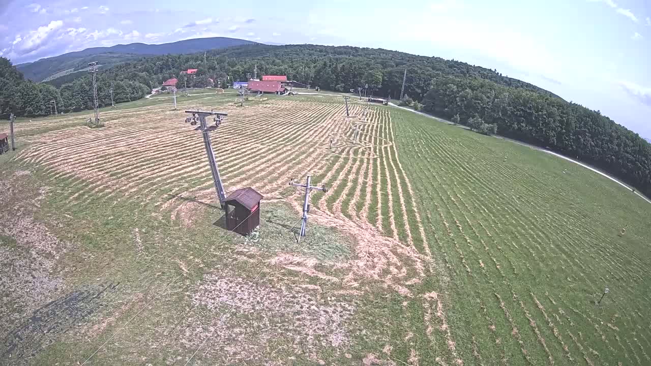 A grassy field with parallel lines cut into it, two ski lifts, and a small building, is situated below a line of trees and mountains under a partly cloudy sky.