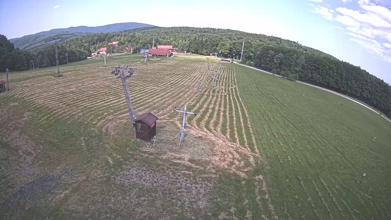 An aerial view shows a grassy field with parallel lines cut into it, ski lift equipment, and a few small buildings in the background under partly cloudy skies.