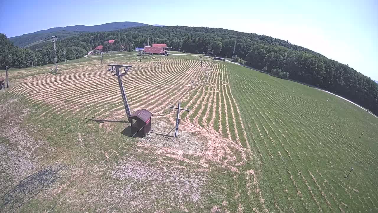 A mowed grassy field with ski lift infrastructure is visible under a clear, sunny sky, surrounded by a forested mountain range.