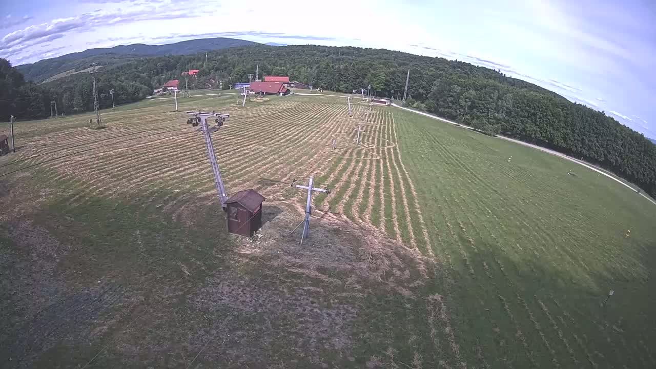 An aerial view shows a grassy field with ski lift infrastructure, a small building, and a forested mountain range in the background under partly cloudy skies.