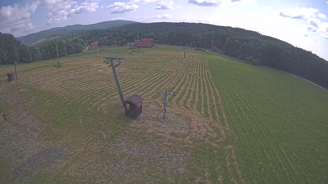 A large, mowed field with two ski lift poles and a small wooden structure is visible beneath a partly cloudy sky, bordered by trees and mountains in the distance.