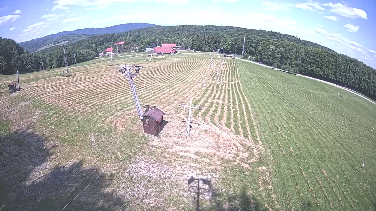 A grassy field with a ski lift infrastructure is visible under a partly cloudy sky, surrounded by a forest-covered mountain range.