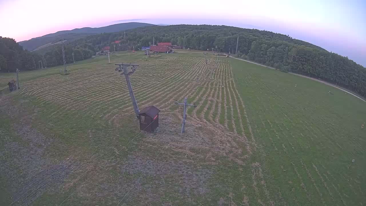 A grassy field with a ski lift infrastructure is visible under a partly cloudy sky, surrounded by a forest-covered mountain range.