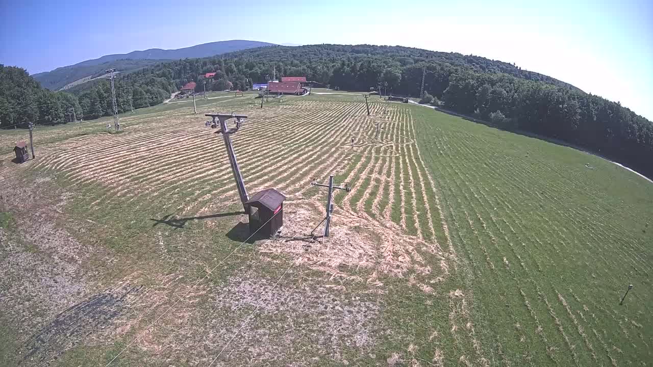 A grassy field with parallel lines cut into it, ski lift infrastructure, and a small building, all situated against a backdrop of forested hills under a clear blue sky.