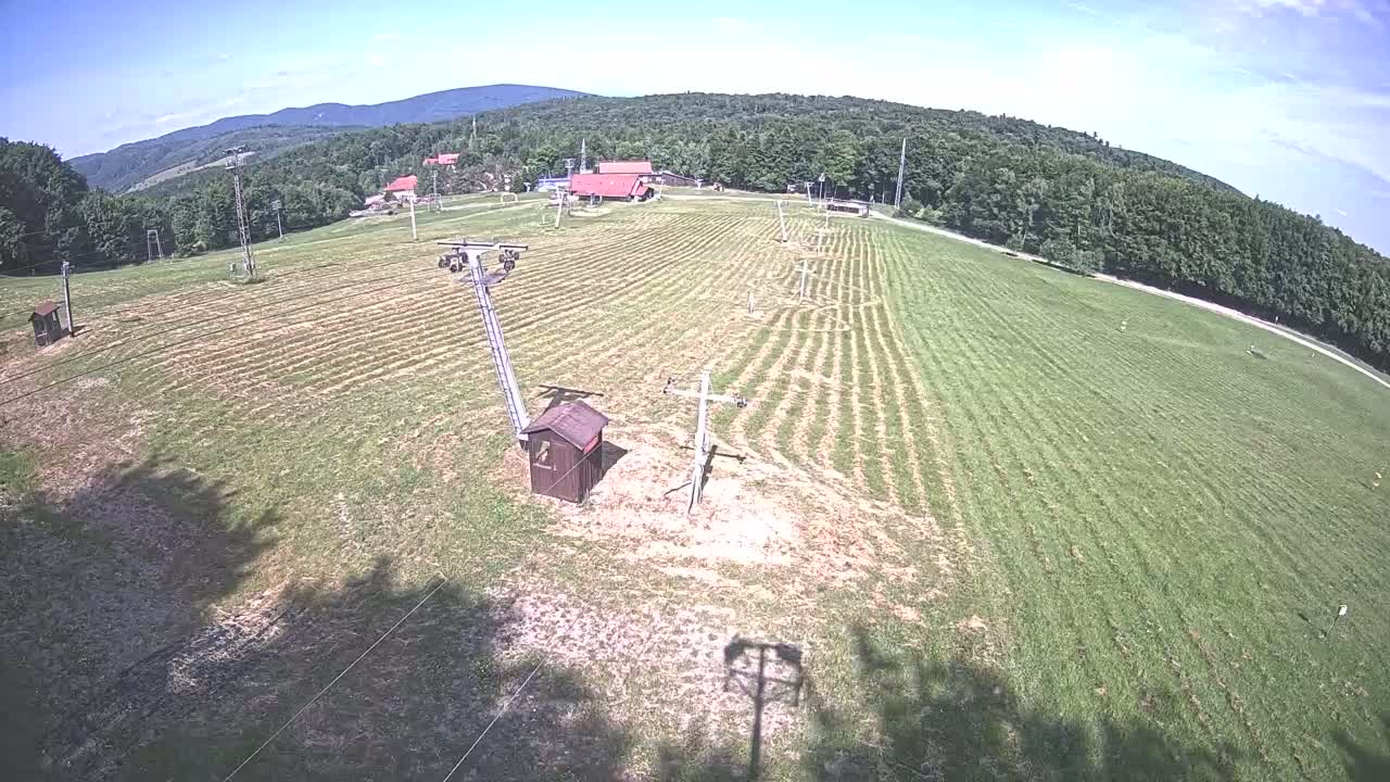 A grassy field with ski lift infrastructure, a small building, and distant forested hills under a clear, sunny sky.