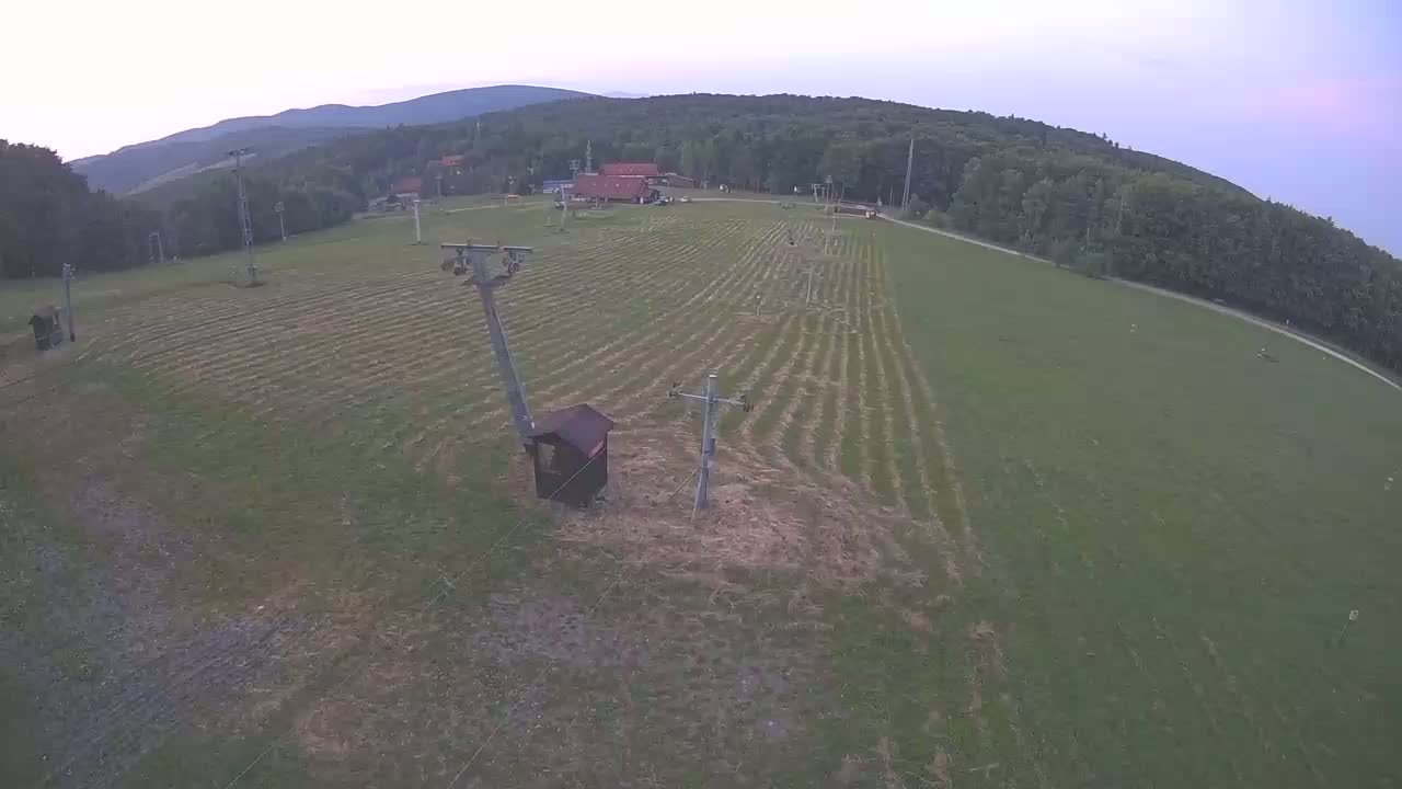 An aerial view shows a grassy field with several ski lift poles and a small building, surrounded by a forest under a dusky sky.