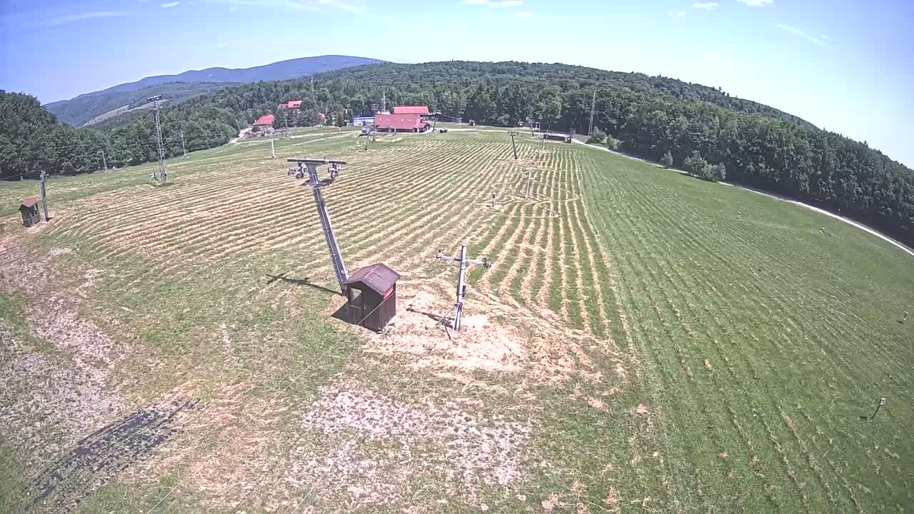 A grassy field with parallel lines cut into it, ski lift infrastructure, and a few buildings in the background under a clear, sunny sky.