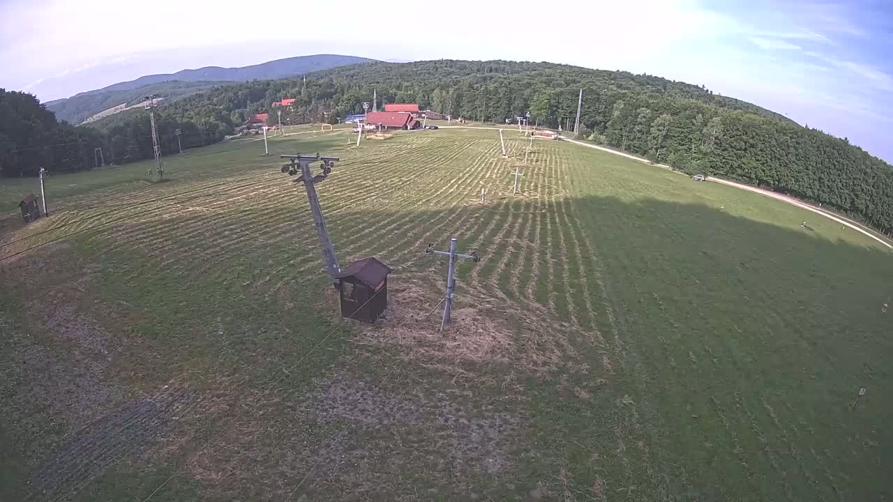 An aerial view shows a grassy field with a ski lift infrastructure, a small building, and a background of a forest and mountains under partly cloudy skies.